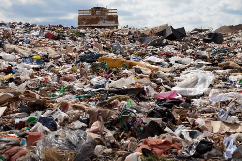 A vast and cluttered landscape of mixed waste materials, including plastic bags, paper, cardboard, and various discarded items, spread across an open area with uneven terrain. In the background, there is a partially constructed or abandoned brick building with scaffolding or structural framing visible on the upper floors. The sky overhead is partly cloudy, casting diffuse light over the scene. The foreground is densely packed with rubbish, emphasizing the scale of waste accumulation, which highlights the importance of professional rubbish removal services such as those provided by House Clearance Bracknell, serving the Bracknell area. The overall environment appears to be an unmanaged waste site or landfill, with debris extending to the horizon and little vegetation visible, underscoring the need for proper waste management and disposal solutions.
