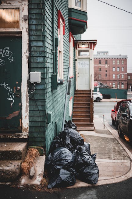 A stack of several black rubbish bags is placed against the exterior wall of a green, wooden-clad building, located on a city street. The bags appear to be filled with household or general waste, tied