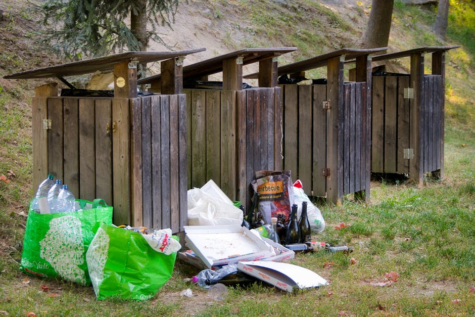 A stack of several black rubbish bags is placed against the exterior wall of a green, wooden-clad building, located on a city street. The bags appear to be filled with household or general waste, tied