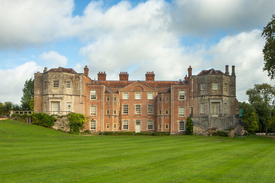 A large historic stone and brick mansion with multiple chimneys and varying window styles, set against a partially cloudy sky. The building features rounded corner towers on both sides with stone deta