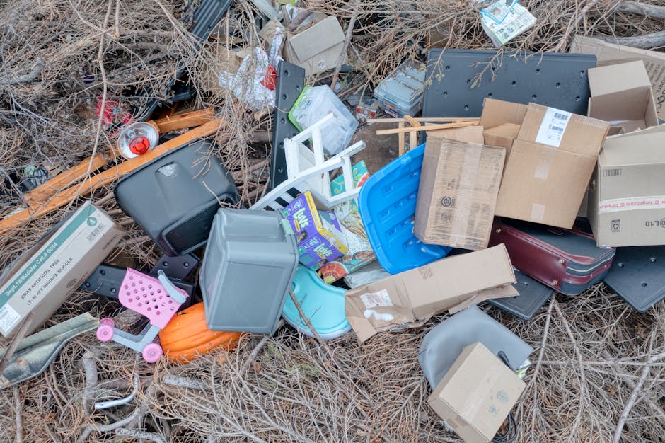 The image depicts a large pile of domestic waste and discarded items scattered across an outdoor setting, likely on a patch of land or overgrown area. Prominently visible at the forefront is a small, 