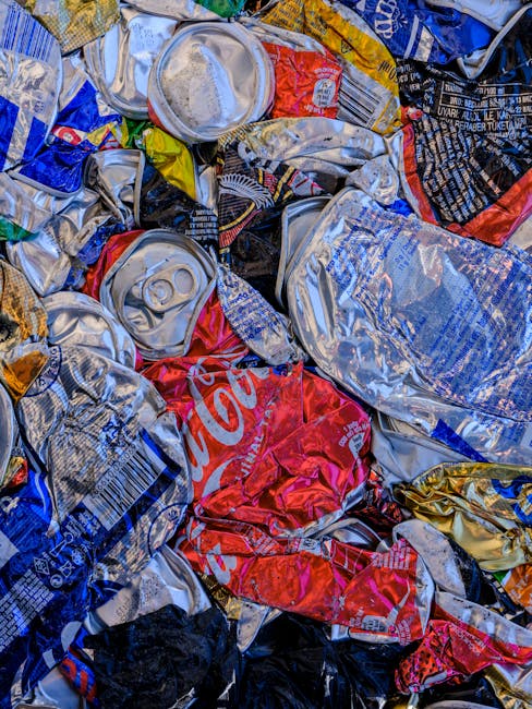 An outdoor scene showing a pile of mixed household waste and discarded items resting on dry, brown grass and leafless branches. The debris consists of various cardboard boxes, some flattened and other